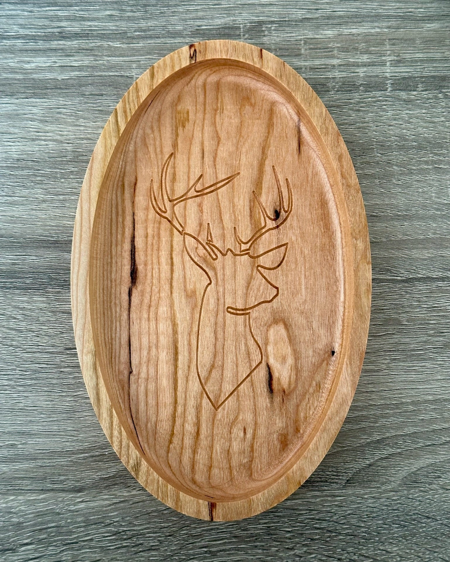 An oval wooden tray with a carved deer head design, displayed on a gray wood-textured background.