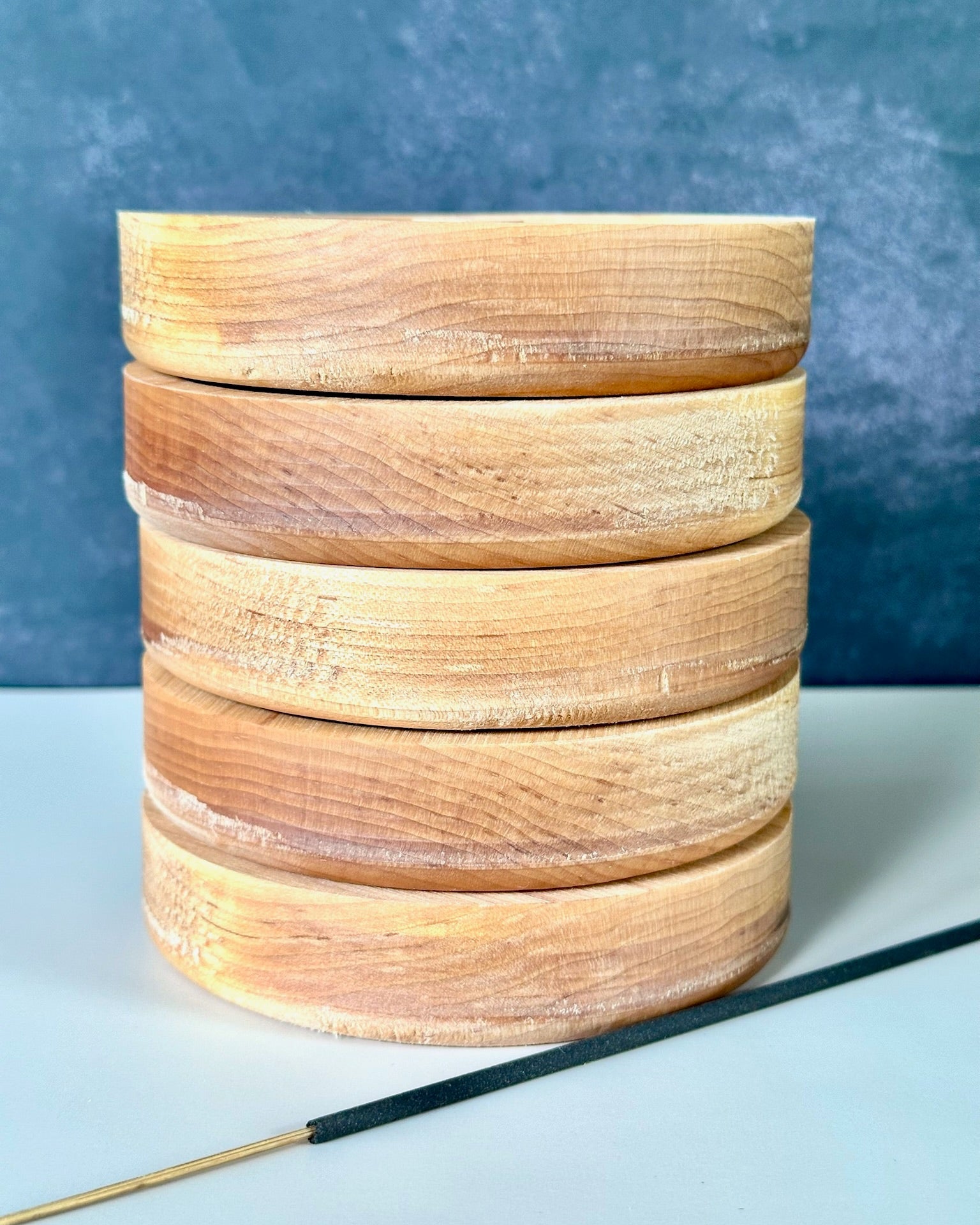 A stack of five round wooden incense burners with visible wood grain, shown on a light surface against a textured blue-gray background, with a black incense stick resting diagonally in the foreground.