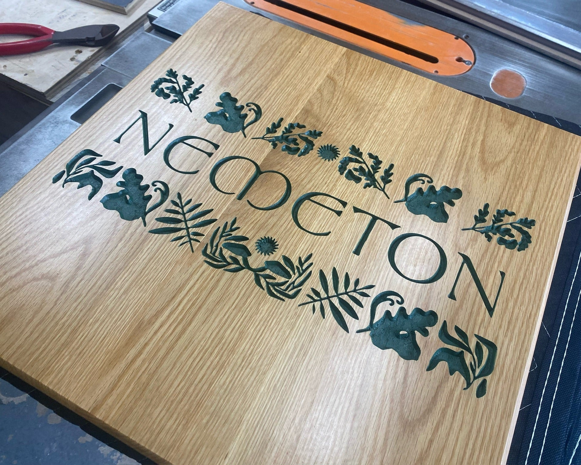 A square wooden oak sign with a natural oak finish, featuring the name 'NEMETON' and decorative floral and leaf motifs in dark green. The board is shown resting on a table saw.
