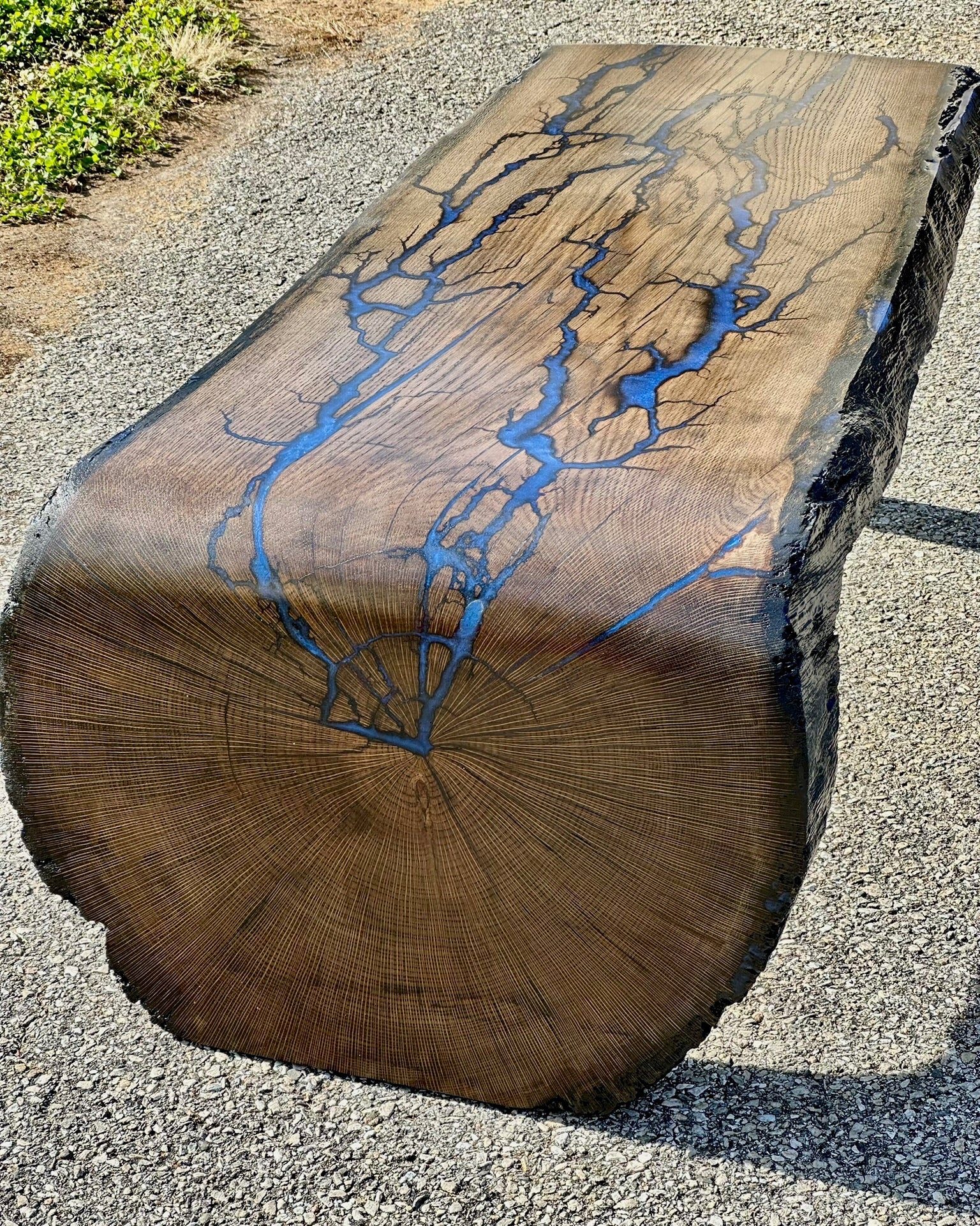 A rustic live-edge wooden bench made from a natural tree log, featuring a prominent blue resin fractal burns running through the wood grain, photographed outdoors on a gravel surface.