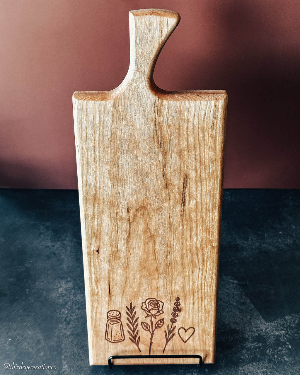 A rectangular wooden cutting board with a handle, standing on a small black metal stand. The board has a natural light wood grain and is decorated with engraved dark brown illustrations of a salt shaker, a rose, lavender sprigs, and a heart at the bottom.