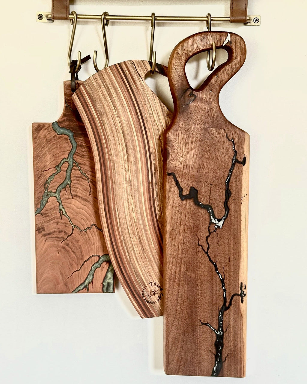 Three wooden serving boards with unique grain patterns and inlay details hanging from metal hooks on a wooden rack against a plain wall.