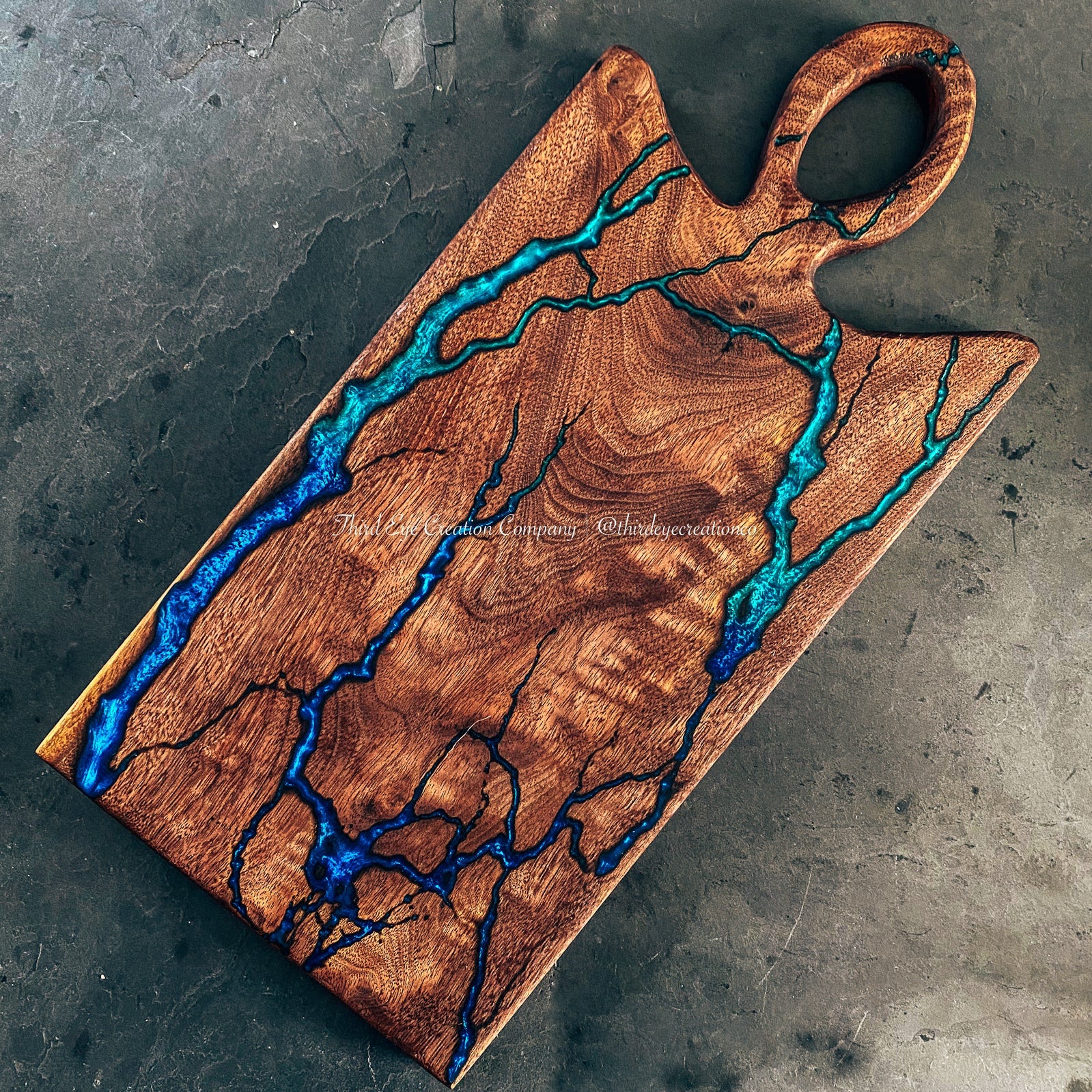 A rectangular wooden cutting board with a loop handle, featuring dramatic natural wood grain and bright blue and turquoise resin fractal burned inlay, photographed from above on a textured gray surface.