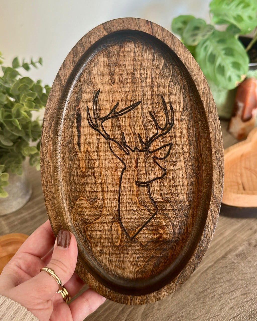 An oval wooden serving tray with a carved deer head design, held by a hand in a cozy indoor setting with plants in the background.