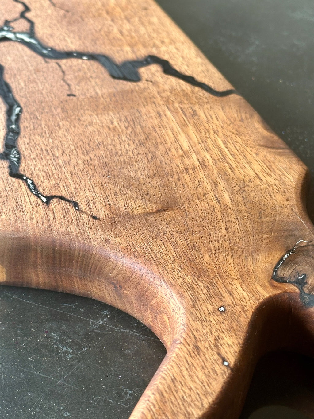 Close-up of a wooden board  with a rich medium-brown wood grain and dark black resin inlay running through the surface, set against a dark gray background.