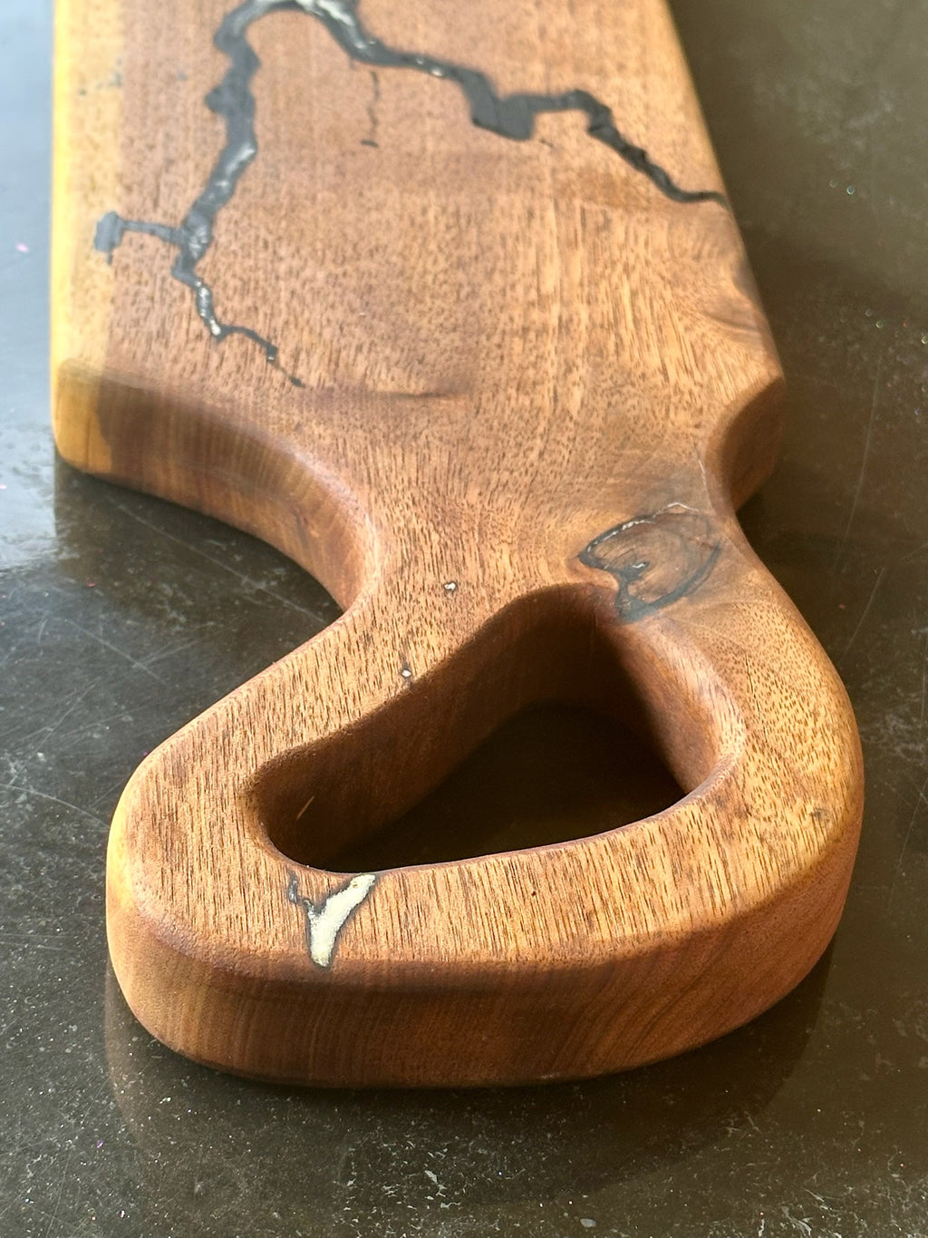 A close-up of a handcrafted wooden cutting board with a heart-shaped handle and dark resin inlay, resting on a dark stone surface.