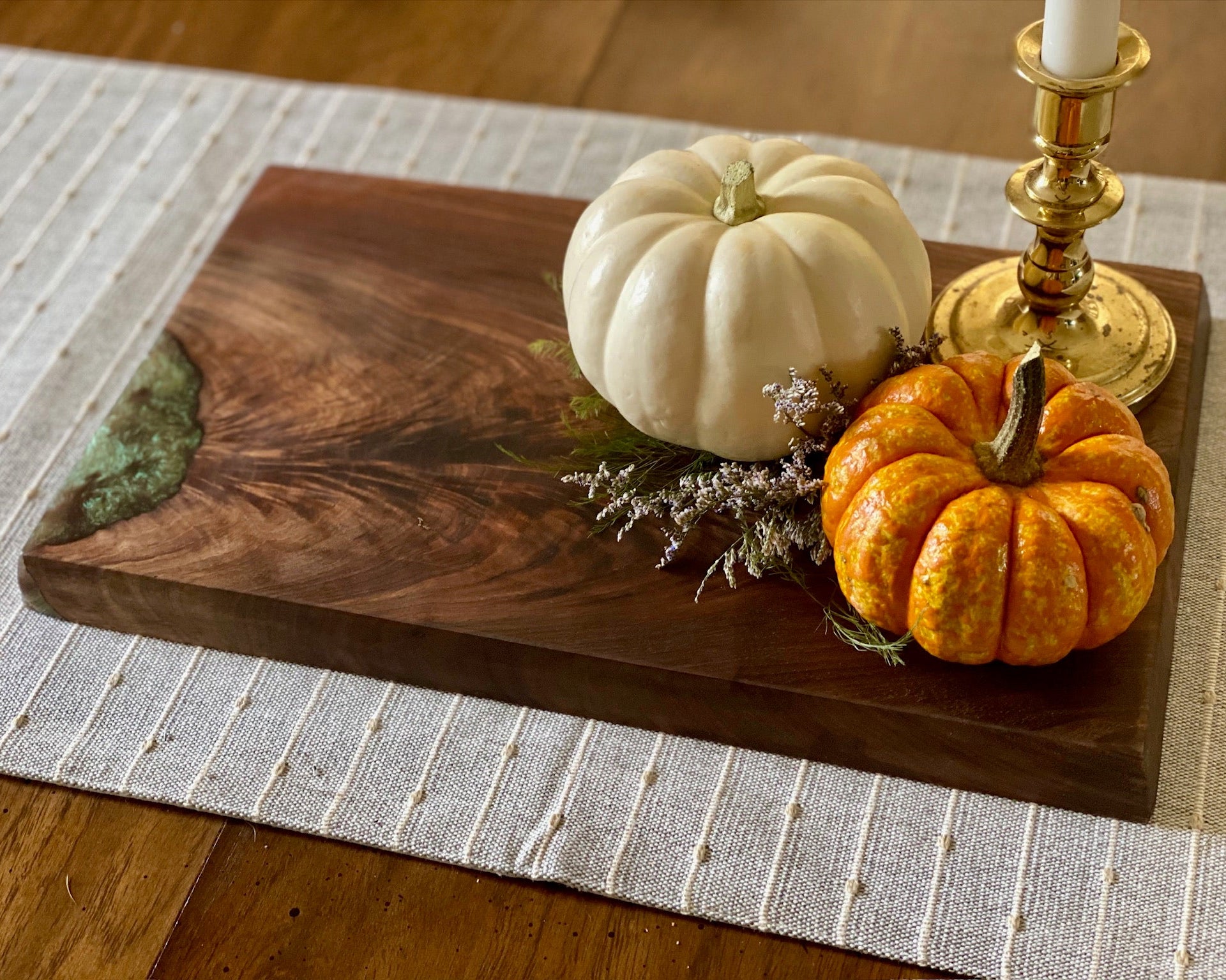 A dark brown wooden serving board with a green resin inlay, styled with a white pumpkin, a small orange pumpkin, dried lavender, and a gold candlestick on a woven placemat over a wooden table.