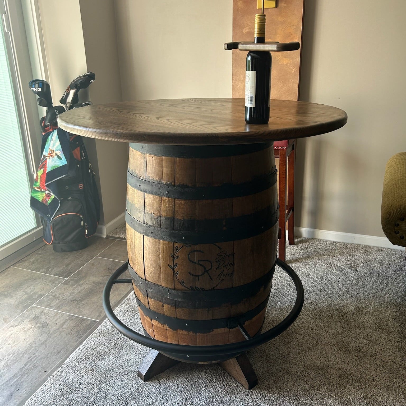 A rustic home bar table made from a repurposed wooden whiskey barrel, featuring a round stained wood top, dark metal reinforcing bands, a circular black metal footrest ring, and a small black pedestal base. A wine bottle is placed on top, and the table is set in a residential room with a gray carpet, beige walls, and a golf bag in the background.