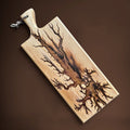 A rectangular wooden cutting board with a handle and a small leather hanging loop, featuring a dramatic tree branch resin design burned into the surface. The board is photographed at an angle against a solid dark brown background.