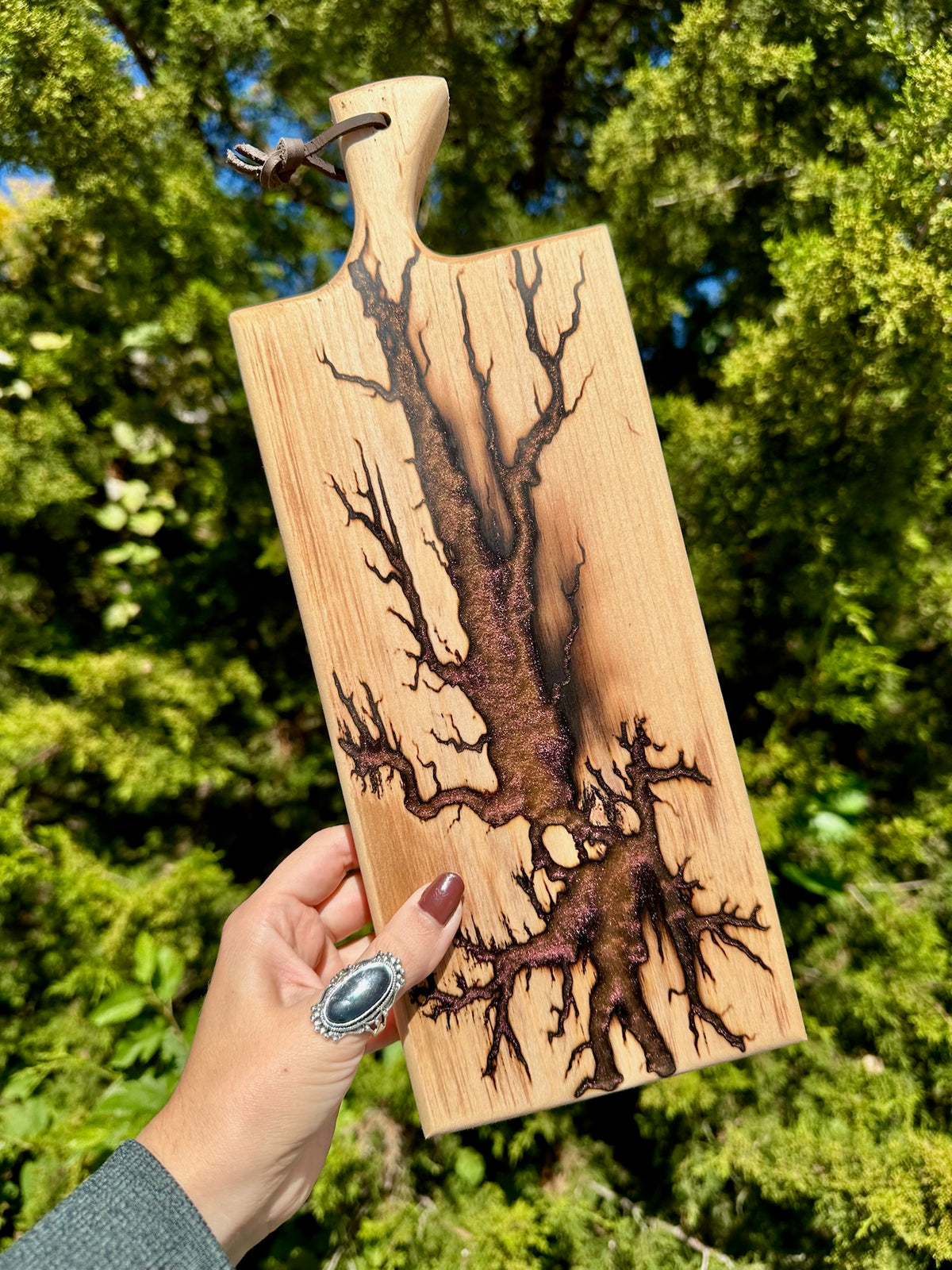 A hand holds a rectangular wooden cutting board with a natural wood grain and dark charred burn patterns resembling tree roots, set against a background of green foliage.
