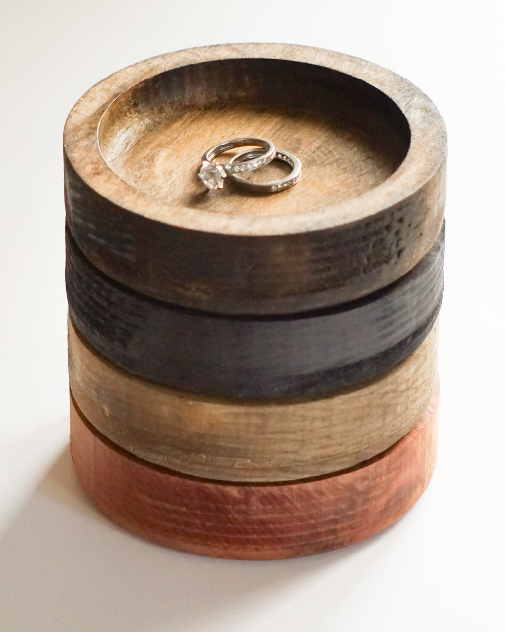 A stack of four round wooden ring dishes in varying natural wood tones, with two silver rings displayed on the top stand, set against a plain white background.