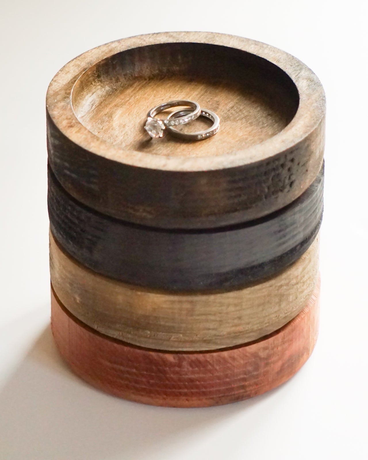 A stack of four round wooden ring dishes in varying natural wood tones, with two silver rings displayed on the top stand, set against a plain white background.